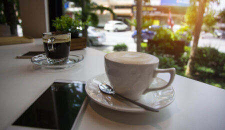 A cup of cappuccino with a spoon on a saucer sits on a table next to a smartphone and a small glass of black coffee. A window reveals a view of greenery and parked cars outside in daylight.