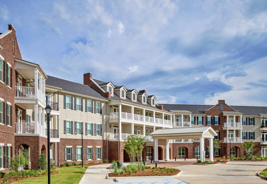 Three-story brick and beige building with balconies, white pillars, and a covered entrance—ideal for downsizing your home after 55—surrounded by landscaped greenery under a partly cloudy sky. A wheelchair-accessible parking space is visible in front.