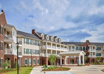 Three-story brick and beige building with balconies, white pillars, and a covered entrance—ideal for downsizing your home after 55—surrounded by landscaped greenery under a partly cloudy sky. A wheelchair-accessible parking space is visible in front.