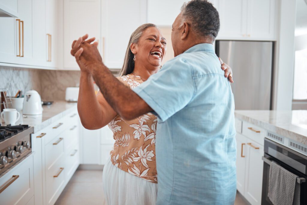 An older couple smiles and dances together in a bright, modern kitchen, enjoying each other's company. The woman in a floral blouse and the man in a light blue shirt experience the many benefits of dancing as they move with joy.