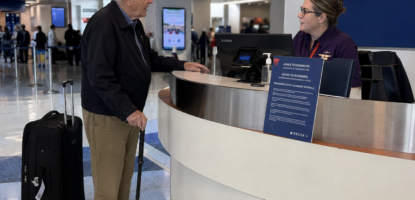 An older man with a cane and rolling suitcase talks to a Delta airline employee at an airport counter, staying alert to travel scams. The employee is behind a desk with a computer; signs for ticketing, check-in, and restrooms are visible in the background.