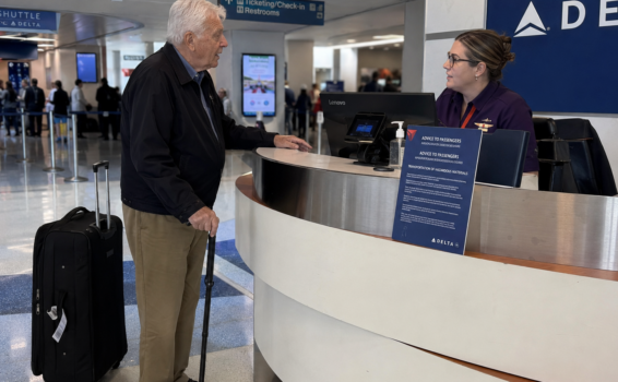 An older man with a cane and rolling suitcase talks to a Delta airline employee at an airport counter, staying alert to travel scams. The employee is behind a desk with a computer; signs for ticketing, check-in, and restrooms are visible in the background.