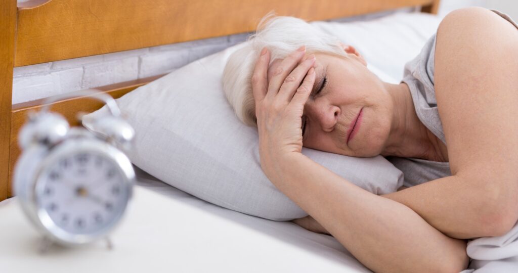 An older woman with short gray hair lies in bed, holding her forehead as if in discomfort, reminding us of the importance of healthy habits after 60. A blurred alarm clock sits on the bedside table in the foreground.