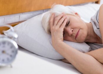 An older woman with short gray hair lies in bed, holding her forehead as if in discomfort, reminding us of the importance of healthy habits after 60. A blurred alarm clock sits on the bedside table in the foreground.