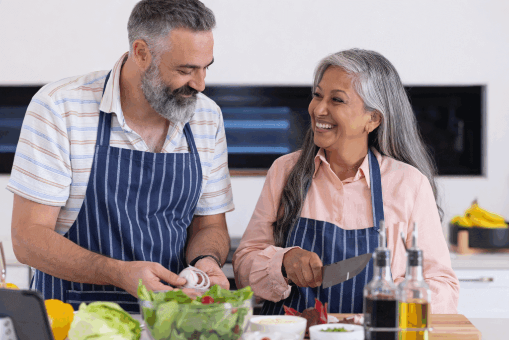 An older man and woman, both wearing striped aprons, smile while preparing vegetables together in a kitchen using safe kitchen tools for seniors. Salad ingredients, oil bottles, and fresh produce are visible on the counter in front of them.