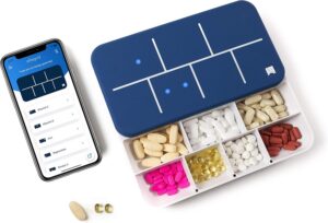 A blue and white pill organizer with seven compartments, each filled with different pills, sits next to a smartphone showing a medication tracking app—one of the top Amazon wellness finds. Two loose capsules rest near the phone.