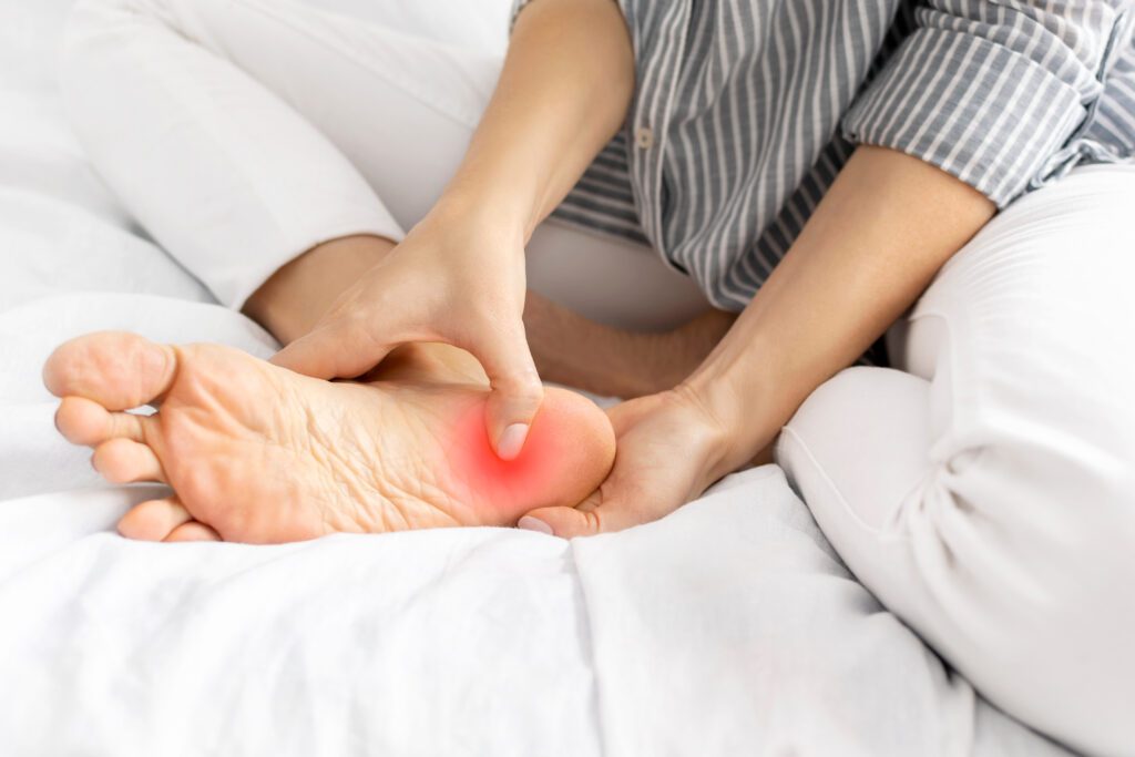 A person sitting on a bed and holding their bare foot, pressing on the heel, which is highlighted with a red area to indicate pain or discomfort.