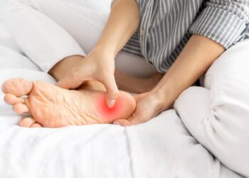 A person sitting on a bed and holding their bare foot, pressing on the heel, which is highlighted with a red area to indicate pain or discomfort.