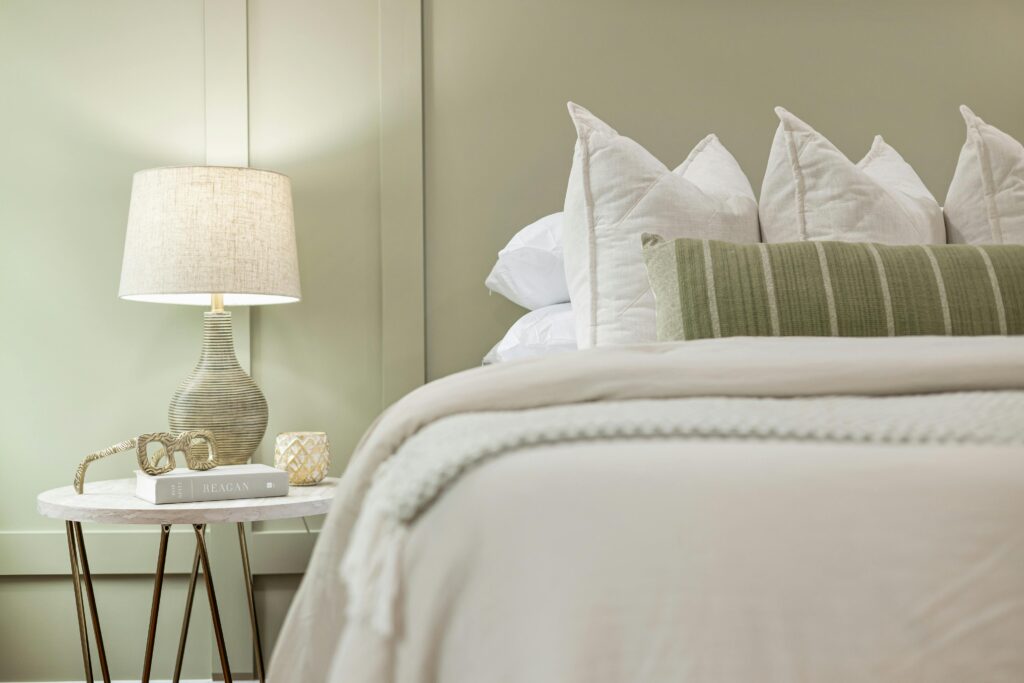 A neatly made bed with white linens, white and green accent pillows, and a beige throw blanket showcases current bedroom design trends. A bedside table holds a textured lamp, a candle, eyeglasses, and books against a light green wall.