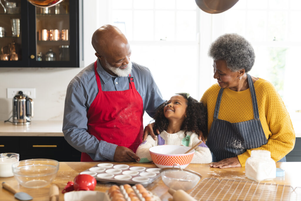 An older man and woman smile at a young girl in a kitchen. The man wears a red apron, the woman a yellow sweater, and the girl stands by a mixing bowl—ready to enjoy easy meals to cook with grandkids amid baking supplies on the wooden counter.