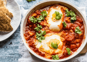 A skillet of shakshuka with poached eggs in a spiced tomato sauce, topped with fresh cilantro. Pita bread is served on a plate beside the skillet.