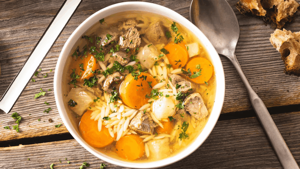 A bowl of hearty German beef soup with chunks of meat, carrots, pasta, and herbs sits on a wooden table next to a spoon, knife, and slices of bread.