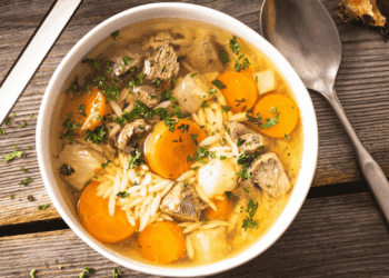A bowl of hearty German beef soup with chunks of meat, carrots, pasta, and herbs sits on a wooden table next to a spoon, knife, and slices of bread.