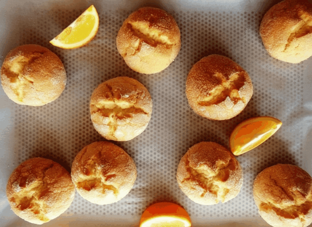 Ten golden brown rounds of grandma’s Turkish citrus cookies with cracked tops are arranged on a baking tray lined with parchment paper, accompanied by three orange wedges placed between the cookies.