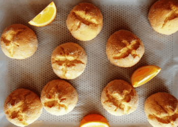Ten golden brown rounds of grandma’s Turkish citrus cookies with cracked tops are arranged on a baking tray lined with parchment paper, accompanied by three orange wedges placed between the cookies.