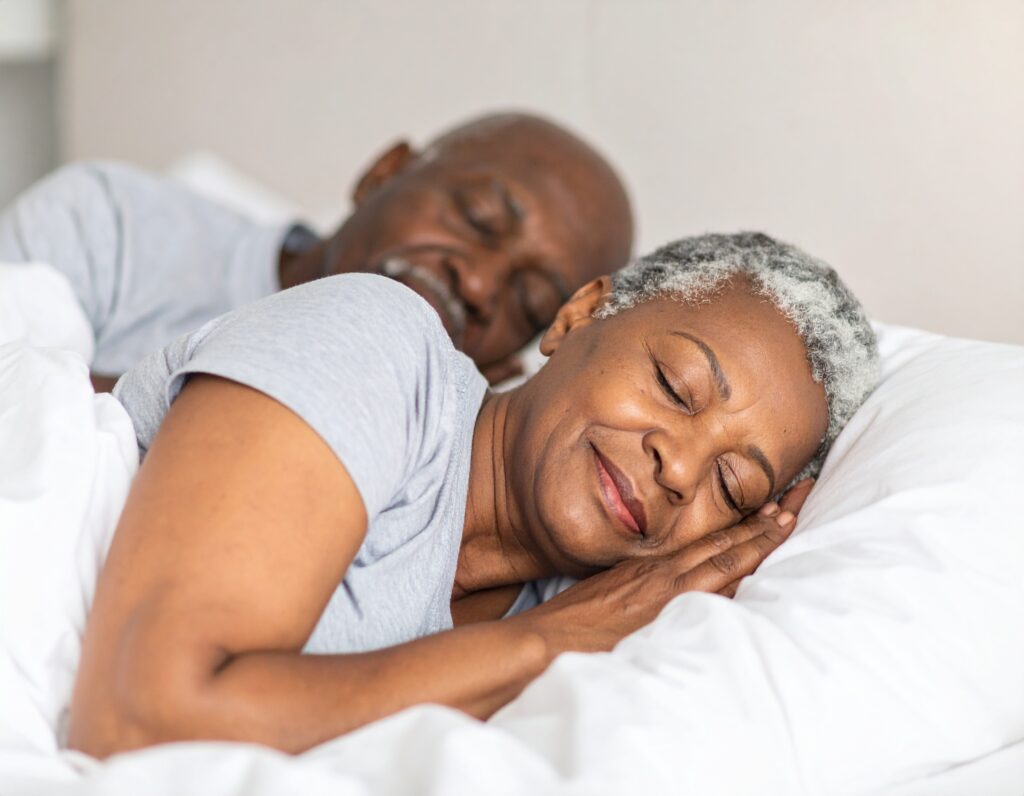 An older couple with gray hair lies in bed, sleeping peacefully under white blankets. Both wear light gray shirts and appear relaxed and content, enjoying restful sleep that supports a healthy metabolism.