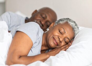 An older couple with gray hair lies in bed, sleeping peacefully under white blankets. Both wear light gray shirts and appear relaxed and content, enjoying restful sleep that supports a healthy metabolism.