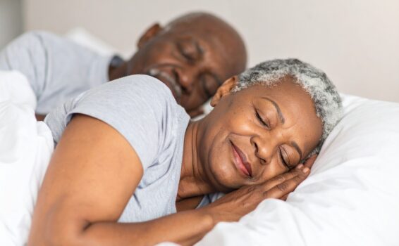 An older couple with gray hair lies in bed, sleeping peacefully under white blankets. Both wear light gray shirts and appear relaxed and content, enjoying restful sleep that supports a healthy metabolism.