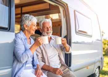 An older couple sits by their camper van, smiling and holding mugs, enjoying a sunny day outdoors. The van door is open, greenery surrounds them—a perfect moment capturing the joy of RV travel.