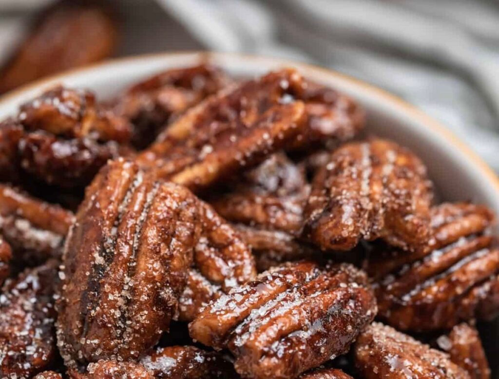 Close-up of a bowl filled with citrus maple candied pecans sprinkled with sugar, showcasing their glossy, textured surfaces and rich brown color.