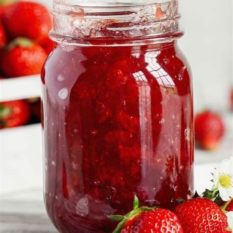 Close-up of a mason jar filled with vibrant homemade strawberry jam, with fresh strawberries in the background.