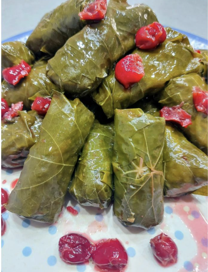 A close-up of neatly rolled grape leaves stuffed with rice, garnished with bright red sour cherries, served on a white plate with blue polka dots. The leaves are glossy and tightly packed.