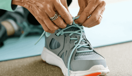 Close-up of a person’s hands tying light blue laces on a gray and white athletic shoe—one of the best shoes for seniors—with a yoga mat in the background.