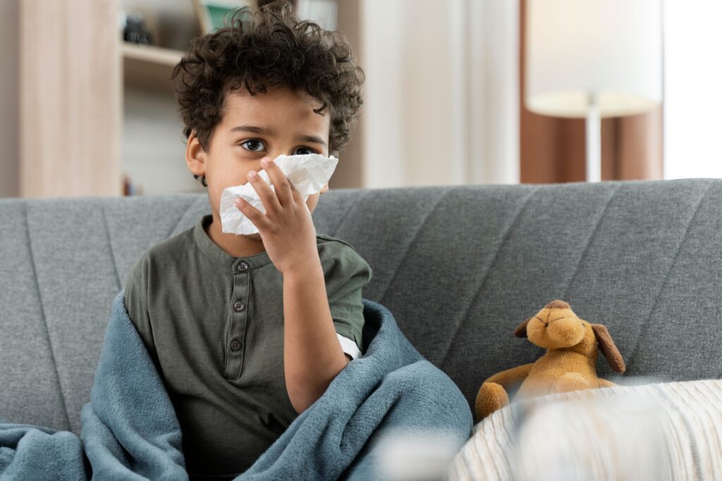 A young child sits on a couch, covered with a blue blanket, holding a tissue to their nose. A brown stuffed animal is next to them as they rest—babysitting your grandkids safely means keeping comfort close when they're feeling unwell.