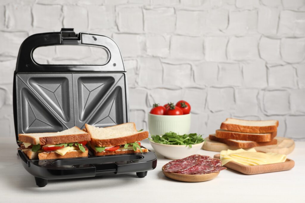 An open sandwich maker, like the best panini press for home use, holds two uncooked sandwiches amid sliced bread, cheese, salami, fresh greens, and tomatoes—all arranged on a white kitchen counter.