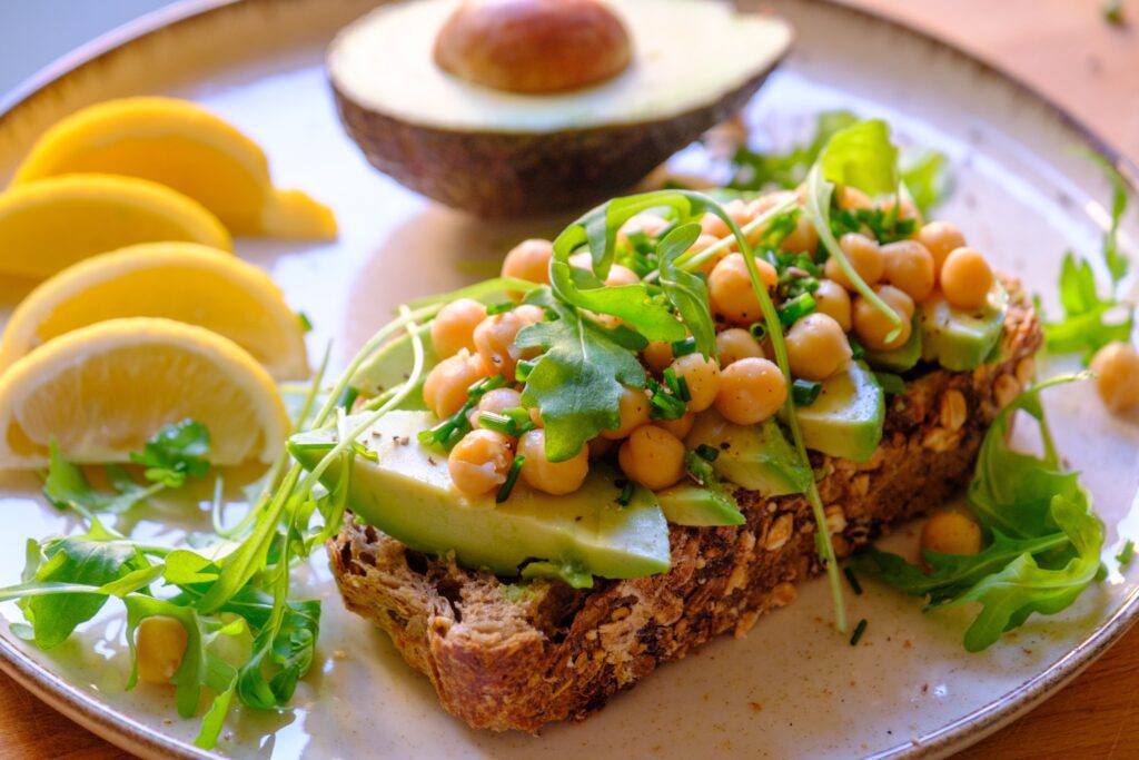 A plate with whole grain bread topped with avocado slices, chickpeas, and arugula highlights fiber benefits for seniors. Beside it are lemon wedges, more arugula, and half an avocado. The dish is fresh and colorful.