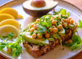 A plate with whole grain bread topped with avocado slices, chickpeas, and arugula highlights fiber benefits for seniors. Beside it are lemon wedges, more arugula, and half an avocado. The dish is fresh and colorful.
