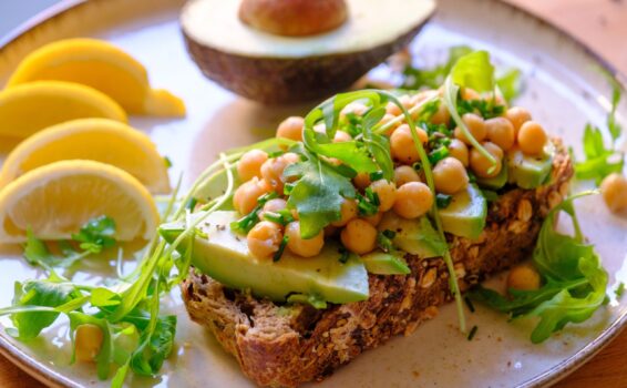 A plate with whole grain bread topped with avocado slices, chickpeas, and arugula highlights fiber benefits for seniors. Beside it are lemon wedges, more arugula, and half an avocado. The dish is fresh and colorful.