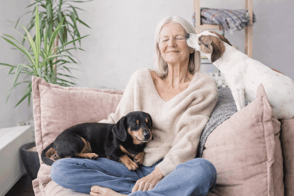 An older woman sits cross-legged on a sofa, smiling with her eyes closed, as one of the best dog breeds for empty nesters rests on her lap and another gently licks her face. A green plant is visible in the background.