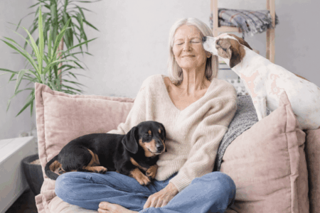 An older woman sits cross-legged on a sofa, smiling with her eyes closed, as one of the best dog breeds for empty nesters rests on her lap and another gently licks her face. A green plant is visible in the background.
