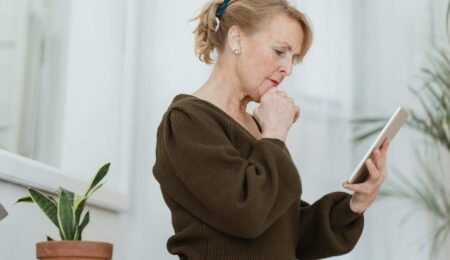 An older woman with blonde hair in a ponytail stands indoors, holding a tablet and looking at it thoughtfully with her hand near her mouth. Nearby, a potted plant adds greenery as she considers easy tech tips for seniors.