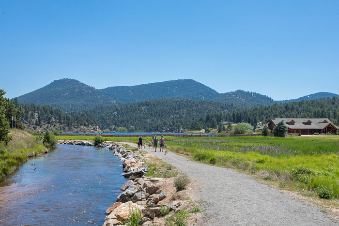 A gravel path runs beside a stream with mountains and trees in the background. People are walking on the path near a lodge, under a clear blue sky—a perfect setting to enjoy outdoor activities in Denver.