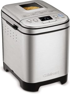 A stainless steel Cuisinart bread maker with a digital display, control buttons, and a viewing window on top, showing a loaf of bread baking inside.
