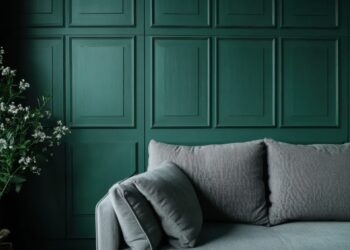A modern room with a gray sofa in front of a dark green paneled wall. A leafy plant with small white flowers is placed on the left side of the image. The scene is minimal and stylish.