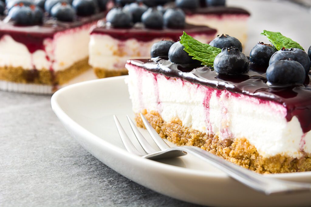 A slice of protein cheesecake with a crumb crust, creamy filling, and blueberry topping is served on a plate with two forks, garnished with fresh blueberries and mint leaves. Other slices are in the background.