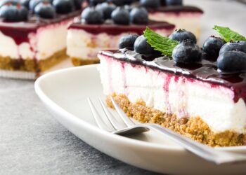 A slice of protein cheesecake with a crumb crust, creamy filling, and blueberry topping is served on a plate with two forks, garnished with fresh blueberries and mint leaves. Other slices are in the background.