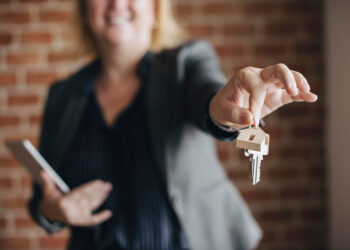 A person in a blazer holds out house keys, smiling, with a brick wall behind them. The focus is on the keys, symbolizing the decision to rent or buy in retirement.