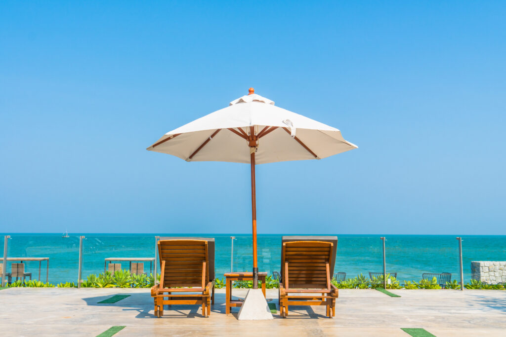 Two wooden lounge chairs sit under a large white umbrella facing the ocean, with a glass railing and clear blue sky in the background on a sunny day.