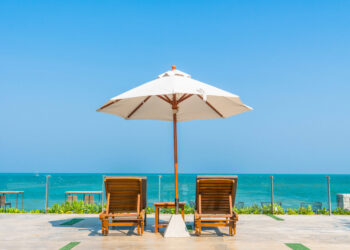 Two wooden lounge chairs sit under a large white umbrella facing the ocean, with a glass railing and clear blue sky in the background on a sunny day.