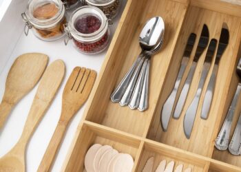 An organized kitchen drawer with wooden utensils, metal spoons and knives in a bamboo divider, and jars of dried food and red peppercorns on the side showcases the benefits of kitchen drawer organizers.