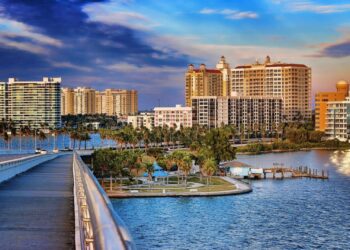 A scenic Sarasota-Florida waterfront cityscape with high-rise buildings, palm trees, a pier, and a park beside blue water, viewed from a bridge under a colorful sky at sunset.