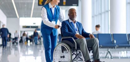 A smiling airport staff member pushes an older man in a wheelchair through a bright, modern terminal, illustrating senior travel safety tips amid glass walls and empty seats in the background.