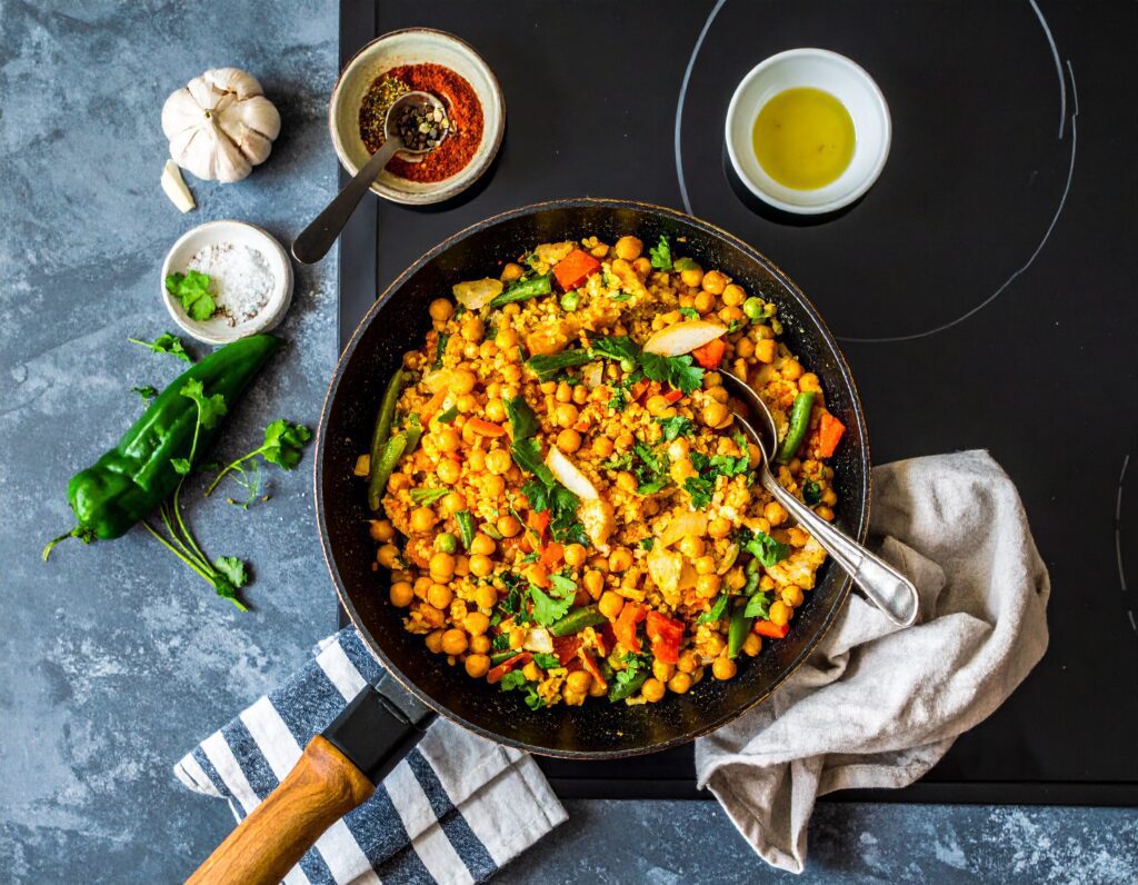 A skillet filled with a colorful plant-based protein skillet meal of chickpea and vegetable stir-fry, garnished with herbs, sits on a striped towel. Nearby are a garlic bulb, green chili, spices, olive oil, and coarse salt on a gray kitchen counter.