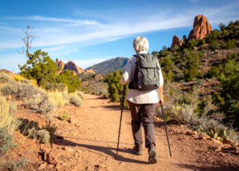 An older adult with gray hair hikes on a dirt trail through a rocky, desert landscape near Denver, enjoying outdoor activities with trekking poles and a backpack, surrounded by shrubs and rock formations under a clear blue sky.
