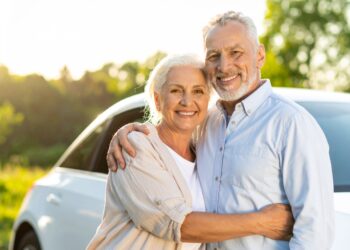 An older couple smiles and embraces in front of a white car outdoors, with green trees and sunlight in the background—enjoying their trip thanks to helpful senior car rental tips.