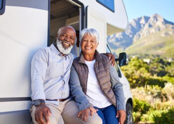 Smiling older couple enjoying RV ownership, sitting together outside their white RV with scenic mountains and greenery in the background on a sunny day.